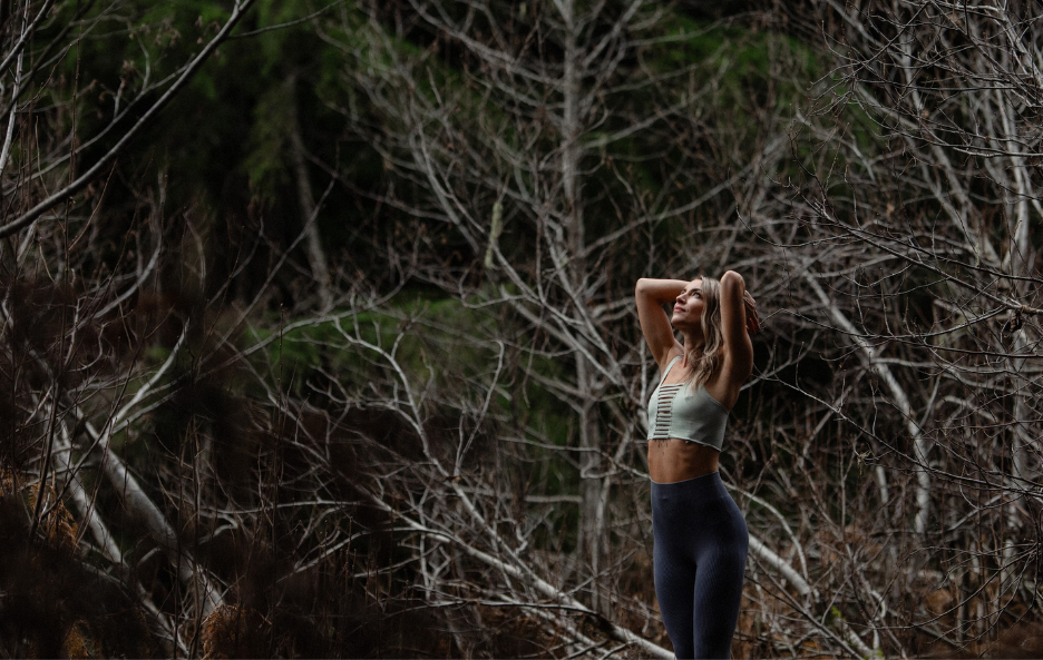 Woman looking up with arms bent at elbow positioned in prayer hands behind her head in wooded forest setting with title reading. Purify. Forge. Triumph.